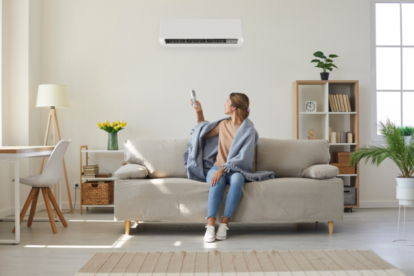 Woman who's sitting on sofa under warm plaid in living room switches off her air conditioner on wall.