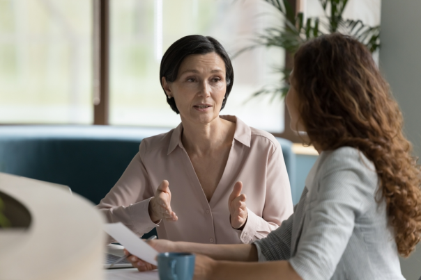 Confident mature business professional woman talking to younger female colleague at office table, speaking, gesturing, teaching, explaining work tasks.